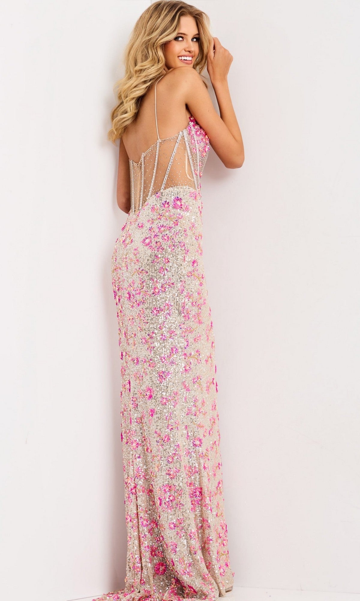 A woman wearing the Jovani 45774 long prom dress with pink and silver beaded embellishments stands facing a white wall, looking over her shoulder and smiling.